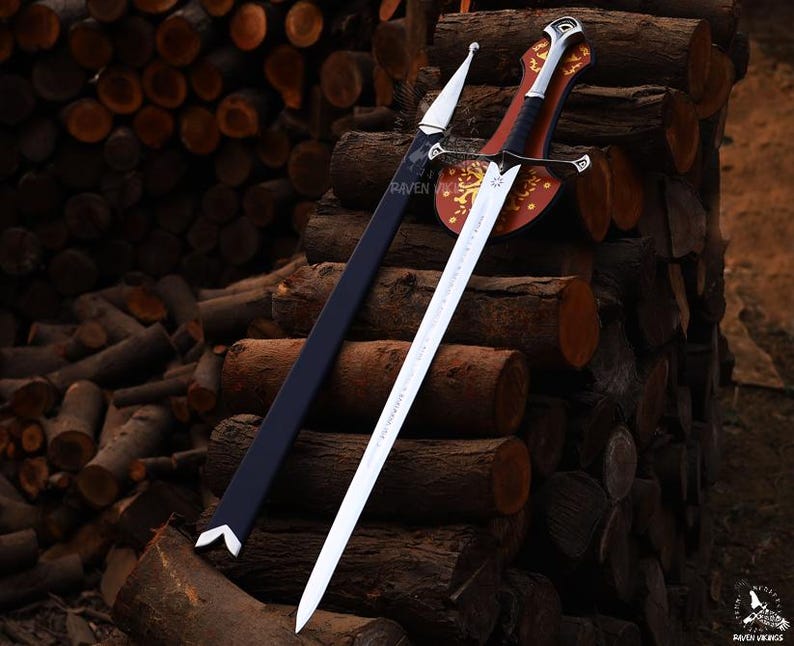 May include: A silver and black sword with a decorative handle and a black sheath. The sword is displayed on a wooden shield with gold accents. The background features a stack of logs. The text "RAVEN VIKINGS" is visible.