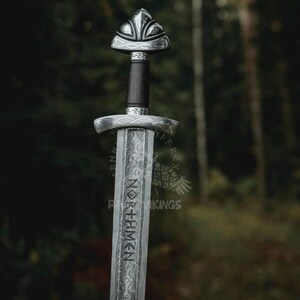 May include: A detailed, silver-colored sword with a dark handle and ornate design. The blade features the word "NORSEMEN" in a vertical inscription. The sword's hilt and pommel are intricately decorated. The background is a blurred forest.