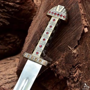 May include: Close-up of a decorative dagger with a silver blade and ornate handle. The handle features gold-colored metal accents and is adorned with red gemstones. The dagger's design suggests a historical or fantasy theme.