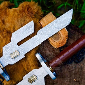 May include: Two silver-colored knives with brown leather handles and a decorative design. The blades have a unique shape with cut-out sections. The handles are wrapped in brown leather. The knives are displayed on a wooden surface with a fur-like background.