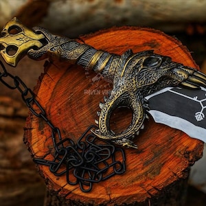 May include: A decorative sword with a detailed gold and bronze handle featuring a dragon design. The blade is black and white with a symbol. A black chain is attached to the handle. The sword rests on a wooden surface.