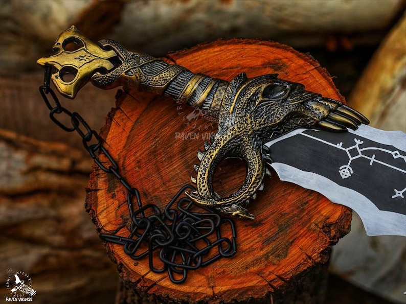 May include: A decorative sword with a detailed gold and bronze handle featuring a dragon design. The blade is black and white with a symbol. A black chain is attached to the handle. The sword rests on a wooden surface.