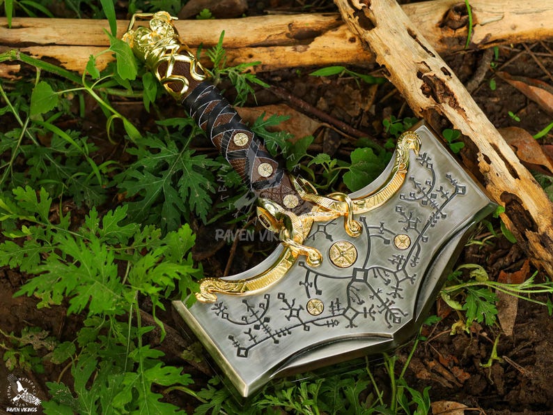 May include: A detailed, metallic Thor's hammer with gold accents and intricate engravings. The hammer features a brown and black leather-wrapped handle and is set against a backdrop of green foliage and wood.