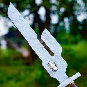 May include: Close-up of a silver metal knife with a unique, angular blade design. The blade has a textured finish and a small, rectangular gold-colored detail. The handle is partially visible, with a brown and gold color scheme.