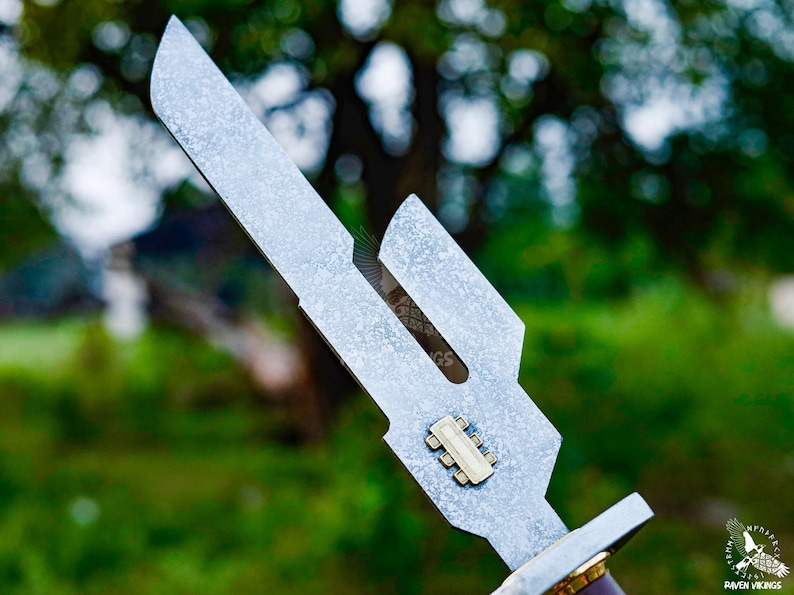 May include: Close-up of a silver metal knife with a unique, angular blade design. The blade has a textured finish and a small, rectangular gold-colored detail. The handle is partially visible, with a brown and gold color scheme.