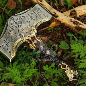 May include: A detailed, metallic hammer with intricate engravings and gold accents. The hammerhead features etched patterns and circular designs. The handle is wrapped in dark leather and gold detailing. The hammer is set against a backdrop of green foliage.