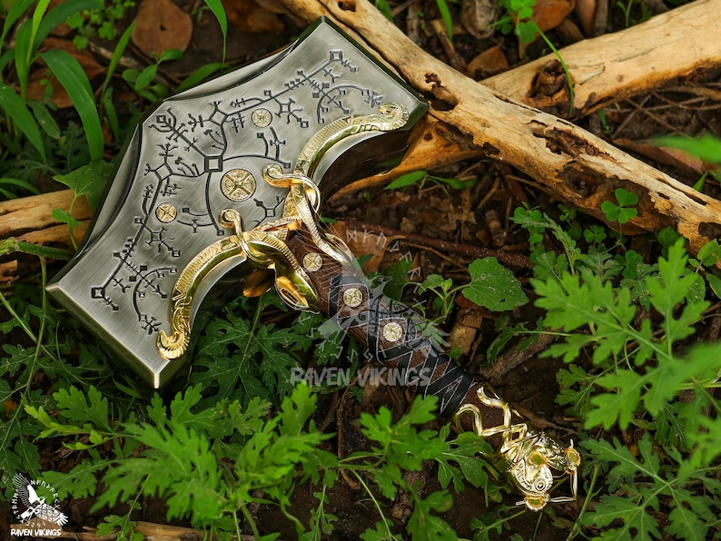May include: A detailed, metallic hammer with intricate engravings and gold accents. The hammerhead features etched patterns and circular designs. The handle is wrapped in dark leather and gold detailing. The hammer is set against a backdrop of green foliage.
