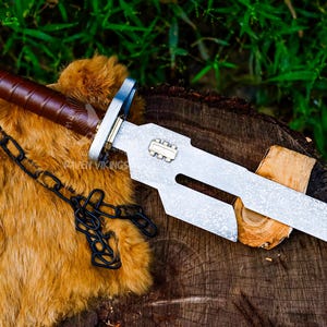 May include: A silver and brown knife with a textured blade and a brown leather-wrapped handle. A black chain is attached to the handle. The knife is resting on a wooden surface with a brown fur-like material.