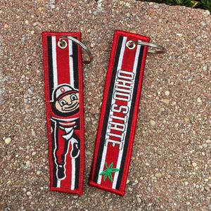 May include: Two rectangular fabric keychains with red, white, and black vertical stripes. One keychain features an embroidered mascot figure, while the other displays the text OHIO STATE in white letters above a small green leaf-like emblem.