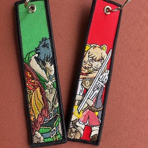 May include: Two embroidered keychains featuring anime characters. The keychain on the left is green with a male character with green hair and a red and orange dragon. The keychain on the right is red with a female character with blonde hair and a sword.