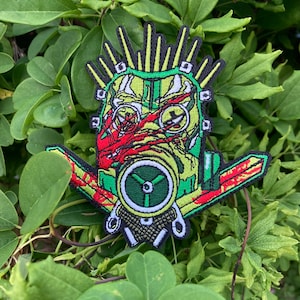May include: Green, red and yellow embroidered patch of a face wearing a gas mask with a mohawk and a large, green, circular fan.
