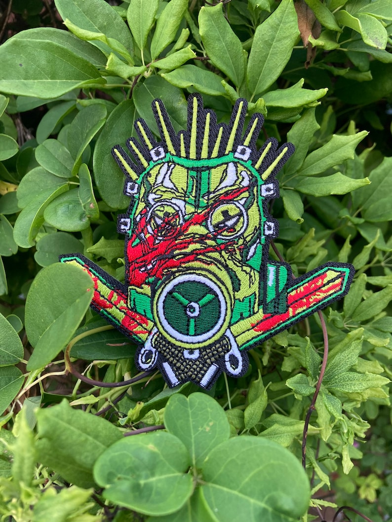 May include: Green, red, and yellow embroidered patch of a face wearing a gas mask with a mohawk and a large, green, circular fan.