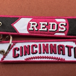 May include: Two red and white fabric keychains with the words "REDS" and "CINCINNATI" embroidered on them. The keychains are attached to a silver key ring.