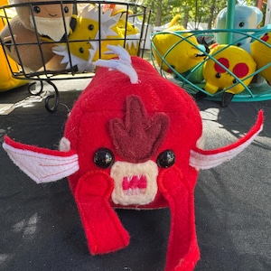 May include: A handmade red plush toy fish with white fins and a dark red design on its head. The toy has black button eyes and a beige mouth. Other plush toys are visible in the background, including a brown fox-like creature and yellow characters.