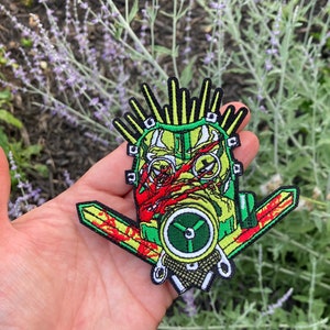 May include: A green, yellow, and red embroidered patch of a face wearing a gas mask with a mohawk and two swords behind the head.