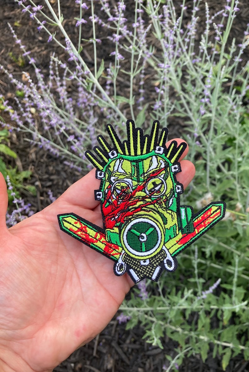 May include: A green, yellow, and red embroidered patch of a face wearing a gas mask with a mohawk and two swords behind the head.