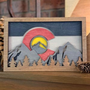 May include: Wooden framed art piece featuring the Colorado state flag and mountain range. The flag design includes a red C shape with a yellow sun, set against a blue, white, and red background. The mountains and trees are gray.
