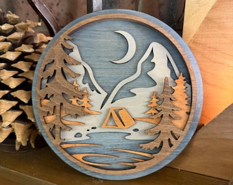 May include: A circular layered wooden wall art piece featuring a mountain landscape with a crescent moon, a tent, a lake, and evergreen trees. The design uses shades of blue, natural wood tones, and cream to create a multi-dimensional scene.