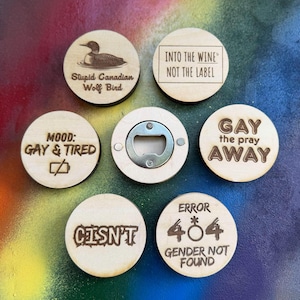 May include: Six circular wooden bottle openers with laser-engraved text and graphics. Designs include a loon with 'Stupid Canadian Wolf Bird', 'INTO THE WINE NOT THE LABEL', 'MOOD: GAY & TIRED', 'GAY the pray AWAY', 'CISN'T', and 'ERROR 404 GENDER NOT FOUND'.