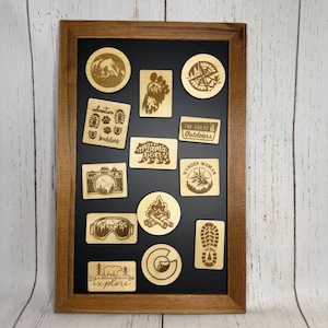 May include: A framed collection of wooden camping-themed magnets. The magnets feature various designs, including a compass, footprints, a camera, and the text "Mama Bear" and "Explore". The frame is a dark brown color.