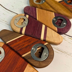 May include: A collection of wooden bottle openers with metal bottle cap removers. The openers feature various wood grains and colors, including reddish-brown, purple, and light brown, with diagonal inlays. The openers are arranged on a light-colored wooden surface.