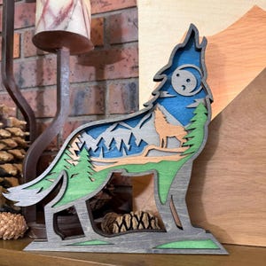 May include: A multi-layered wooden sculpture of a howling wolf. The artwork features a wolf silhouette against a backdrop of mountains, trees, and a moon, with shades of blue, green, and gray. The piece is designed to stand upright.