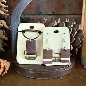 May include: Wooden jewelry set featuring a pendant necklace and earrings. The rectangular pendants and earrings are made of dark wood with a light stripe and a dark purple stripe. The necklace has a leather cord. The earrings have silver hooks. The cards read "Workshop".