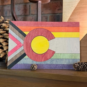 May include: A wooden wall art piece featuring the Colorado state flag design. The artwork incorporates a rainbow of colors, including red, orange, yellow, green, blue, and purple, with a large red "C" and yellow circle. The flag is approximately 12 inches wide and 8 inches tall.