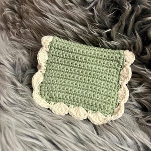 Op de afbeelding: Een handgemaakte gehaakte onderzetter in vierkante vorm. De onderzetter is lichtgroen met een crèmekleurige geschulpte rand. De onderzetter ligt op een grijze, pluizige ondergrond.