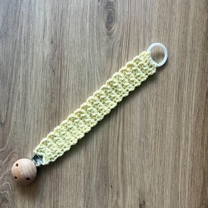 May include: A pale yellow crochet pacifier clip with a wooden bead and a white plastic ring. The clip is made of a textured, woven material and is designed to attach to a pacifier.