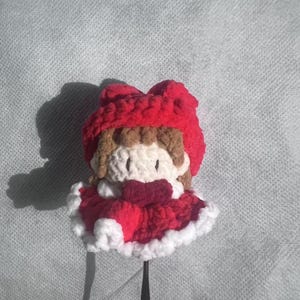 May include: A handmade crochet doll with a red hooded cape and a white-trimmed dress. The doll has brown hair, a white face, and a red bow. The doll is attached to a black wire.