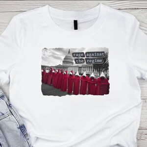 May include: White t-shirt with a black and white image of a group of people wearing red robes and white pointed hats standing in front of the US Capitol building. The text "rage against the regime" is printed in black on a white background.