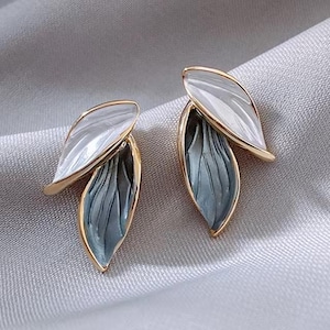 Puede incluir: Un par de pendientes de tono dorado con un diseño en capas. La capa superior es blanca y la capa inferior es azul con un acabado texturizado.