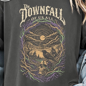 Puede incluir: Camiseta color pimienta con un diseño gótico. El gráfico incluye el texto "The Downfall of Us All" sobre una ilustración detallada de un cuervo, una calavera y un paisaje iluminado por la luna con árboles y un castillo.