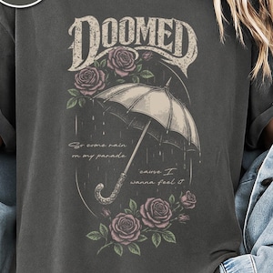 Woman wearing a pepper color Comfort Colors T-shirt featuring a vintage gothic umbrella illustration with roses and rain details and the word “Doomed” in distressed lettering.