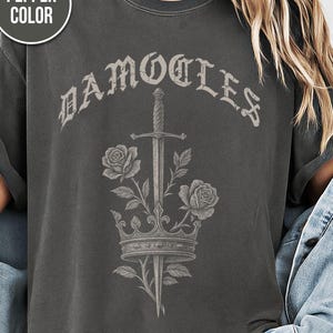 May include: A pepper-coloured t-shirt with a graphic design. The design includes the word "DAMOCLES" in an ornate font, a sword, roses, and a crown. The shirt has a vintage, worn appearance.