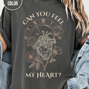 May include: Pepper-colored t-shirt with a graphic design featuring a human heart, roses, and the text "CAN YOU FEEL MY HEART?" The design includes a clock face. The shirt is a dark gray color.