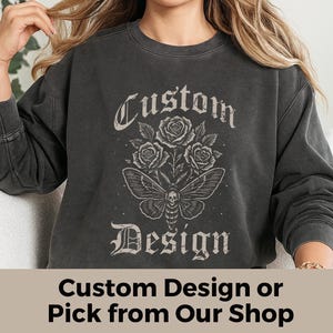 Puede incluir: Sudadera gris oscuro con un diseño personalizado con rosas y una polilla con una calavera en su cuerpo. Las palabras "Custom Design" están impresas encima y debajo del gráfico en una fuente gótica.