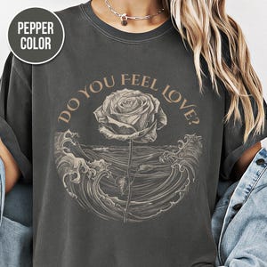 Puede incluir: Camiseta color pimienta con un gráfico de rosa y el texto "Do You Feel Love?". El diseño incluye una rosa que emerge de las olas del océano. La camiseta tiene un aspecto vintage y desgastado.