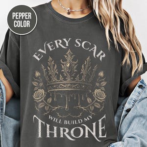 May include: Pepper color t-shirt with a graphic design. The design features a detailed crown with roses and the text "EVERY SCAR WILL BUILD MY THRONE" in a gothic font. The shirt has a vintage, worn look.
