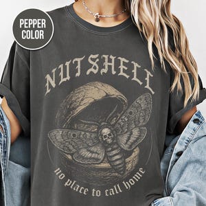 May include: Pepper color t-shirt with a graphic design. The design features the word "Nutshell" above a moth with a skull on its body, emerging from a walnut. The text "no place to call home" is below the moth.