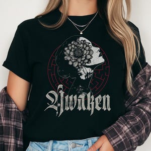Puede incluir: Camiseta negra con una ilustración de un perfil femenino con una flor en el pelo, sobre un fondo de laberinto rojo. La palabra "Awaken" está escrita debajo en fuente gótica. La camiseta se combina con unos vaqueros azules. También se lleva una camisa a cuadros.
