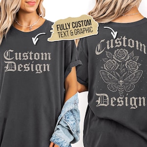 Puede incluir: Una camiseta gris carbón con el texto Custom Design en la parte delantera. La parte trasera muestra un gráfico de tres rosas sobre una polilla con una marca de calavera, acompañado del texto Custom Design. Una etiqueta dice FULLY CUSTOM TEXT & GRAPHIC.