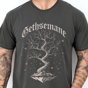 May include: A charcoal gray short-sleeve t-shirt featuring a graphic of a gnarled tree on a floating rock base with small pink blossoms. The word Gethsemane is printed in a distressed, gothic-style font above the tree illustration.