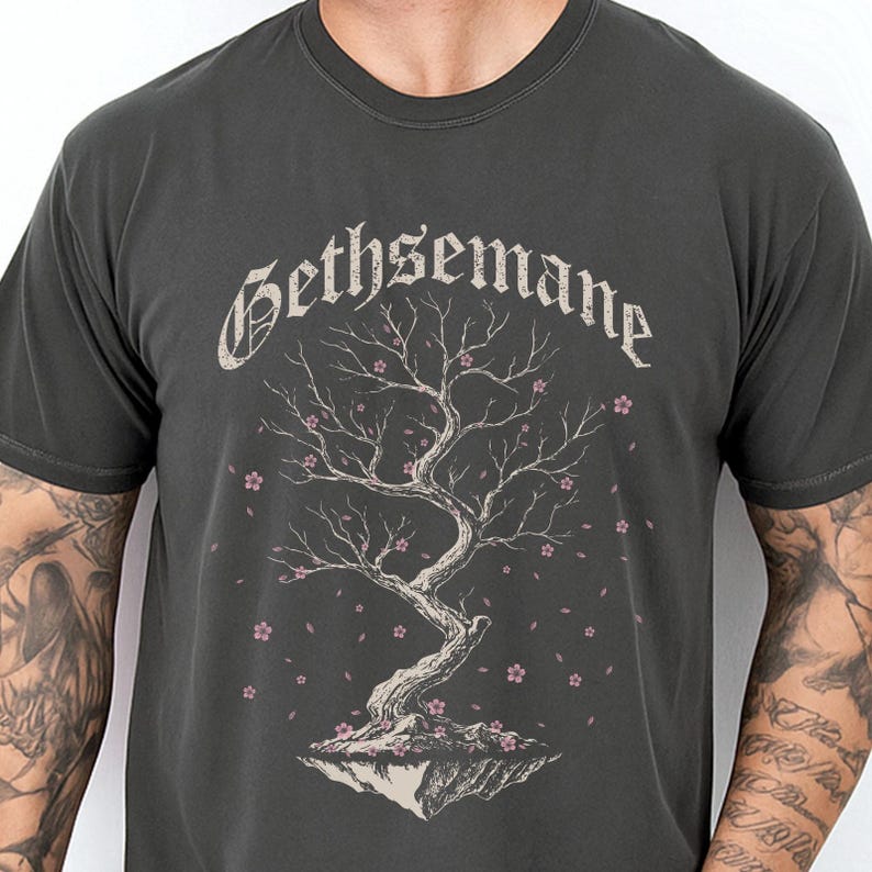 May include: A charcoal gray short-sleeve t-shirt featuring a graphic of a gnarled tree on a floating rock base with small pink blossoms. The word Gethsemane is printed in a distressed, gothic-style font above the tree illustration.