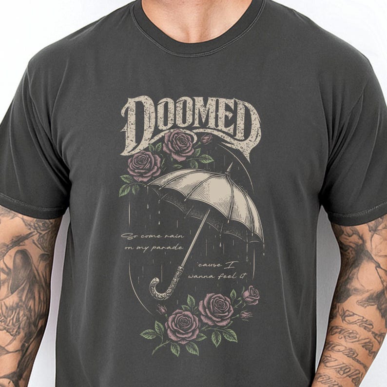 Man wearing a pepper color Comfort Colors T-shirt featuring a vintage gothic umbrella illustration with roses and rain details and the word “Doomed” in distressed lettering.