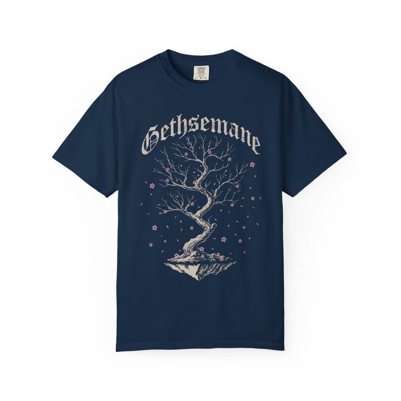 May include: Navy blue t-shirt featuring a tree graphic with the word "Gethsemane" above it. The tree has bare branches and pink blossoms. The design is centered on the shirt.
