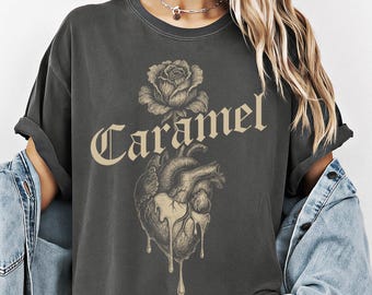 May include: A charcoal gray t-shirt featuring a beige graphic print of an anatomical heart with liquid dripping from the bottom, topped by a rose and the word Caramel in a gothic font. The shirt has short sleeves and a crew neckline.