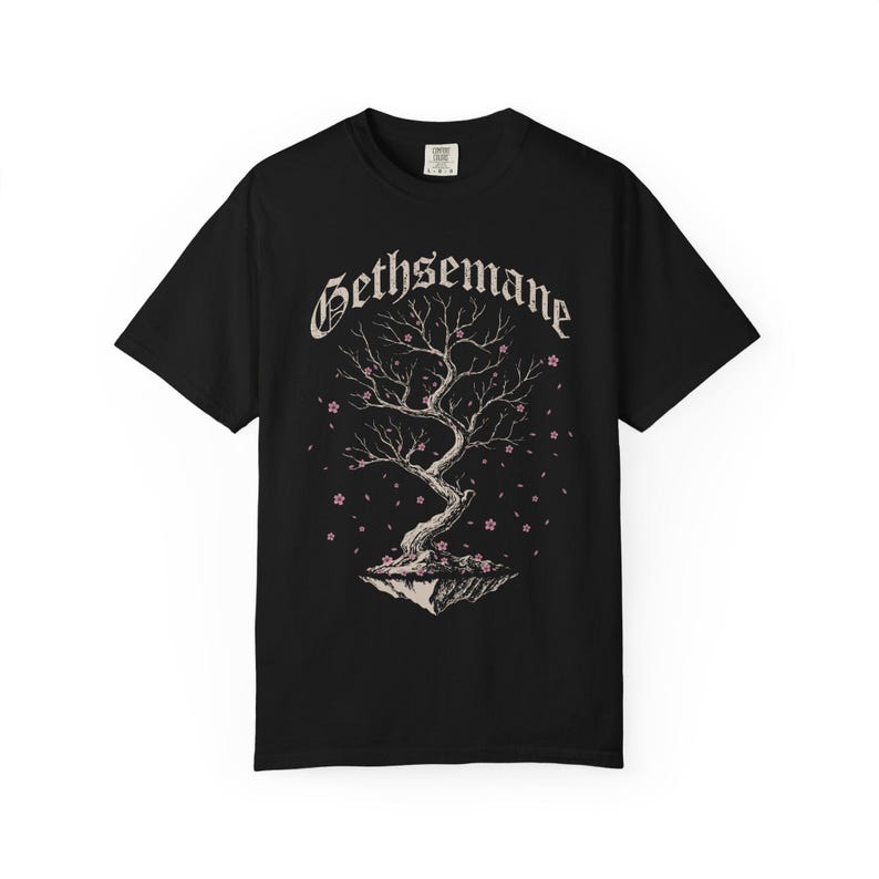 May include: Black t-shirt featuring a stylized tree design with the word "Gethsemane" in an ornate font. The tree has delicate branches and pink blossoms, with some falling around it. The design is centered on the shirt.