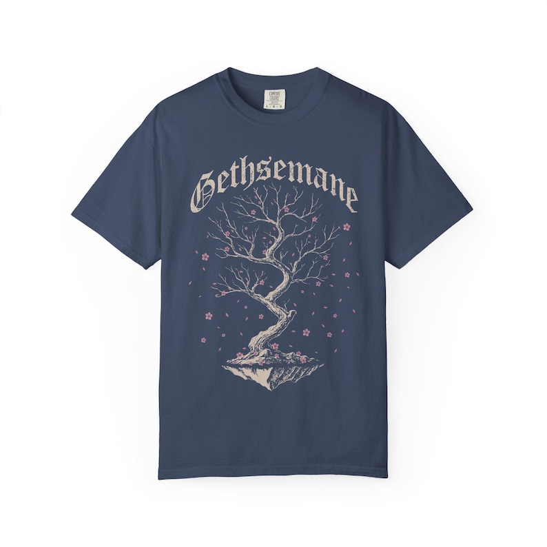 May include: A navy blue short-sleeve t-shirt featuring a graphic of a bare, twisted tree on a floating rock island. The word GETHSEMANE is printed in a white gothic font above the tree, which is surrounded by small, scattered pink flower petals.
